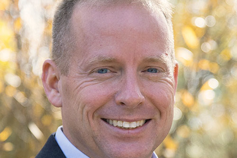 A white man with blue eyes stands outside smiling