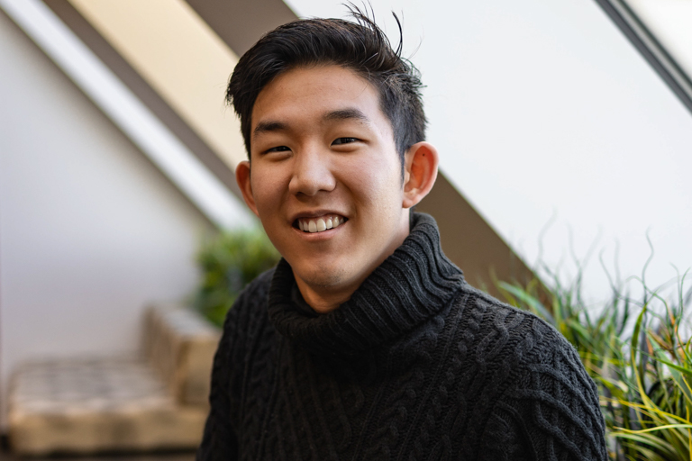 A young East Asian male sitting by a window and wearing a black sweater smiles