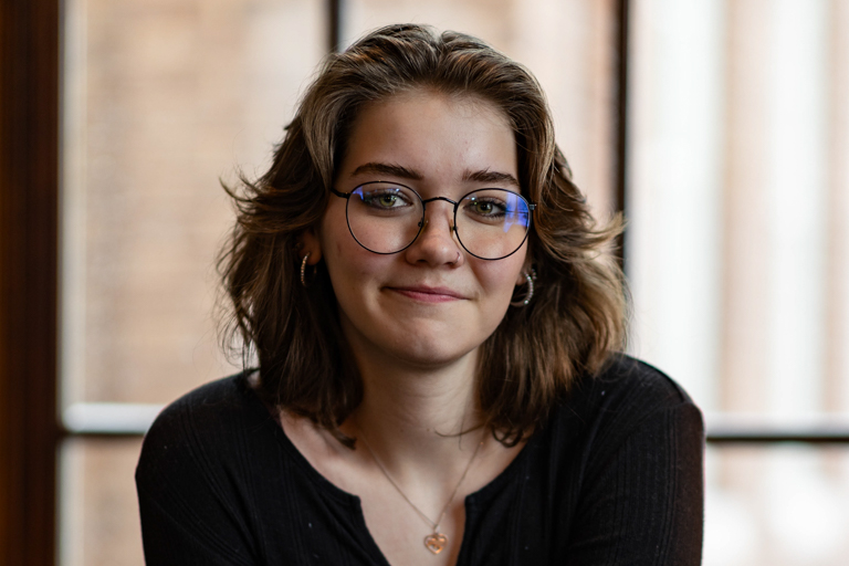 A young white female sitting by a window and wearing a black sweater smiles